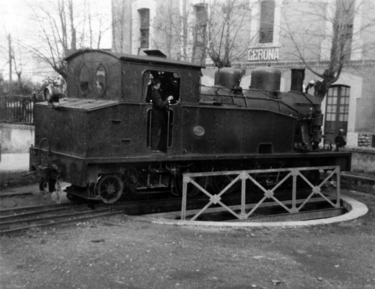 Locomotora de vapor del ferrocarril Olot–Girona davant de l’estació de Girona, amb un ferroviari a la cabina.